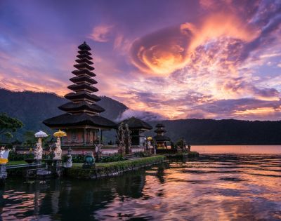 A serene lakeside temple with tiered roofs at sunset in Bali, Indonesia. Vibrant pink and purple clouds reflect on the water, creating a peaceful atmosphere.