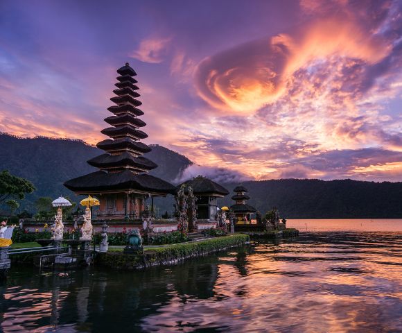 A serene lakeside temple with tiered roofs at sunset in Bali, Indonesia. Vibrant pink and purple clouds reflect on the water, creating a peaceful atmosphere.
