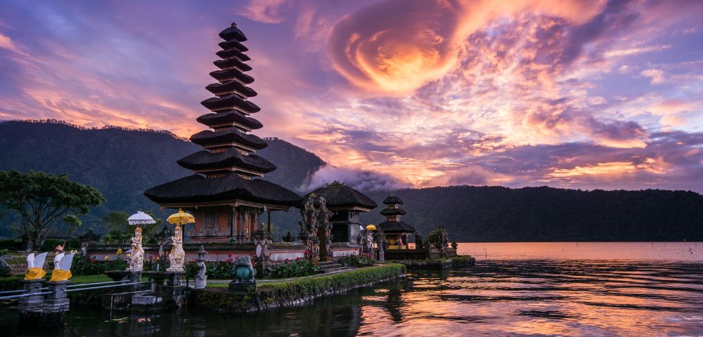 A serene lakeside temple with tiered roofs at sunset in Bali, Indonesia. Vibrant pink and purple clouds reflect on the water, creating a peaceful atmosphere.