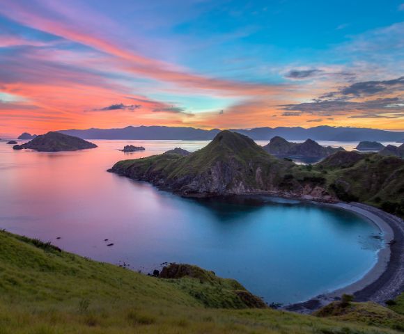 Scenic view of a tranquil bay at sunset, with vibrant pink and blue skies. Hills surround the calm water, creating a serene and peaceful atmosphere.