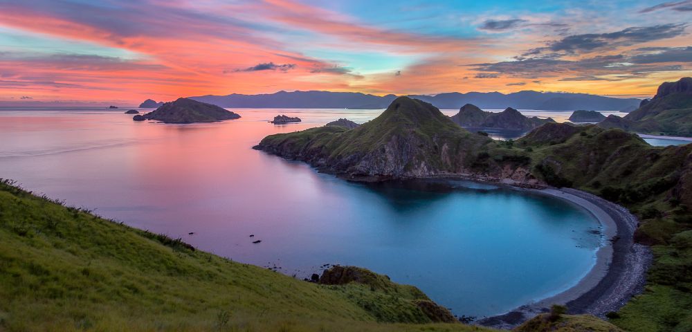 Scenic view of a tranquil bay at sunset, with vibrant pink and blue skies. Hills surround the calm water, creating a serene and peaceful atmosphere.