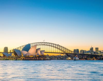 Sydney skyline at dusk featuring the iconic Opera House and Harbour Bridge, with calm waters and a clear, vibrant sky transitioning from gold to blue.