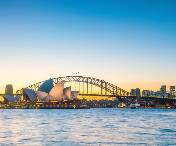 Sydney skyline at dusk featuring the iconic Opera House and Harbour Bridge, with calm waters and a clear, vibrant sky transitioning from gold to blue.