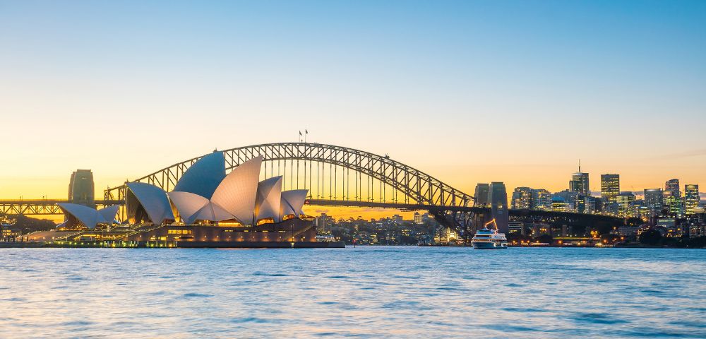 Sydney skyline at dusk featuring the iconic Opera House and Harbour Bridge, with calm waters and a clear, vibrant sky transitioning from gold to blue.