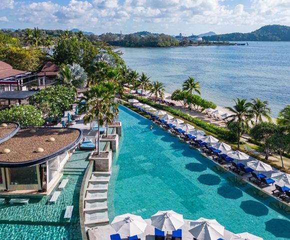 Luxurious beachfront resort with an infinity pool lined by umbrellas and lounge chairs, surrounded by lush greenery, overlooking a serene ocean view.