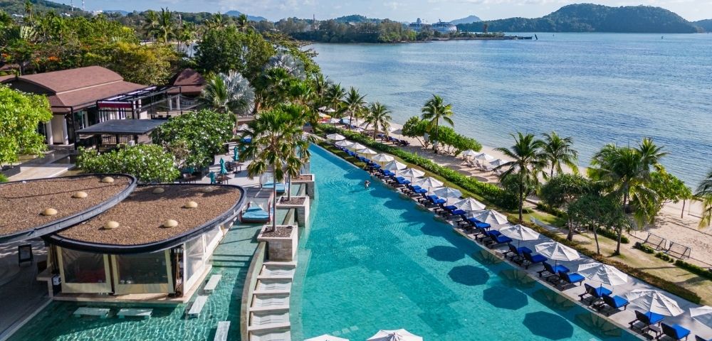 Luxurious beachfront resort with an infinity pool lined by umbrellas and lounge chairs, surrounded by lush greenery, overlooking a serene ocean view.