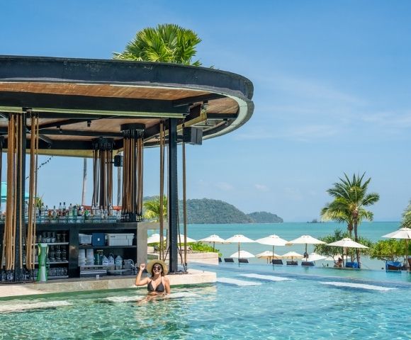 Woman relaxing in a pool near a swim-up bar, with tropical palm trees and ocean views in the background on a sunny day.