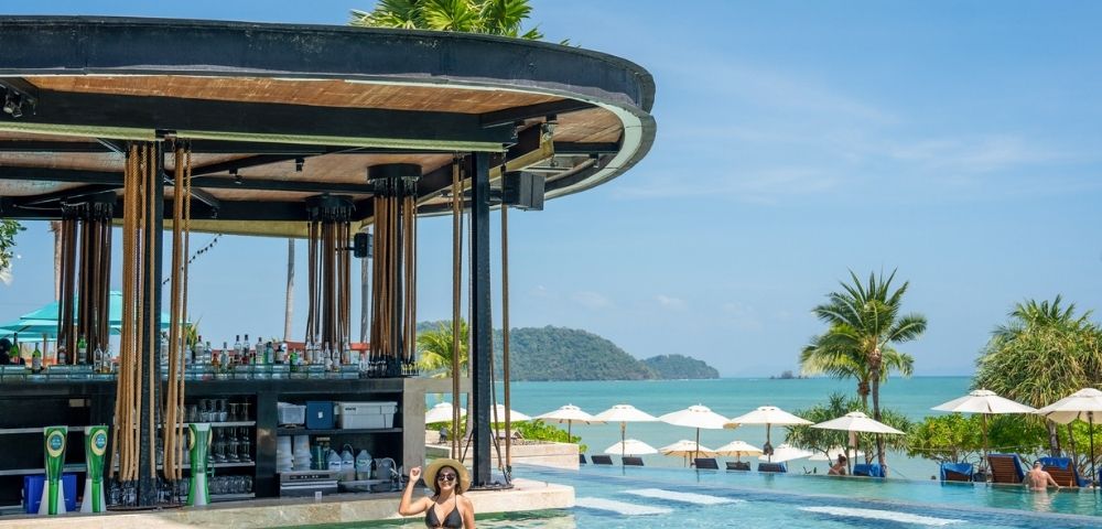 Woman relaxing in a pool near a swim-up bar, with tropical palm trees and ocean views in the background on a sunny day.
