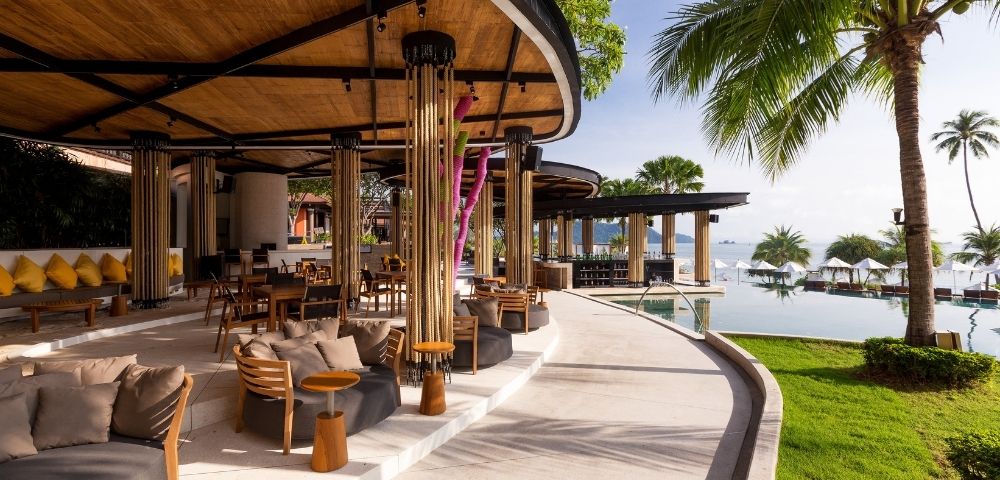 Modern open-air resort lounge with wooden ceiling, cozy seating, and palm trees. Overlooks an infinity pool and ocean under a clear blue sky. Relaxing vibe.