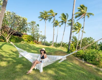 A woman in a sunhat relaxes on a hammock on a lush lawn, surrounded by swaying palm trees under a clear blue sky, conveying a serene tropical vibe.