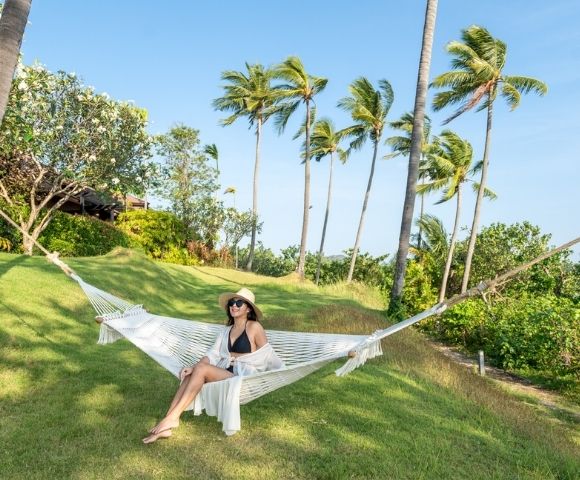 A woman in a sunhat relaxes on a hammock on a lush lawn, surrounded by swaying palm trees under a clear blue sky, conveying a serene tropical vibe.