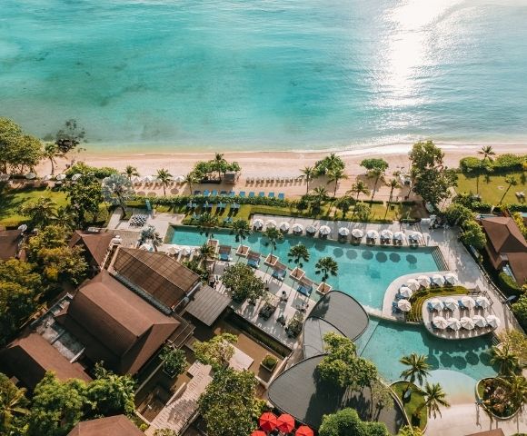Aerial view of a tropical resort with a large pool and surrounding sun loungers, bordered by lush greenery, next to a sandy beach and turquoise ocean.