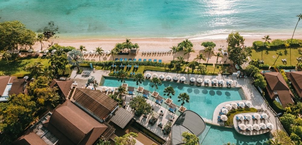 Aerial view of a tropical resort with a large pool and surrounding sun loungers, bordered by lush greenery, next to a sandy beach and turquoise ocean.