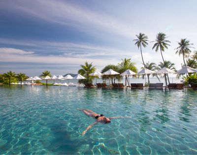 A person floats peacefully in a serene, turquoise pool surrounded by palm trees and white umbrellas under a clear sky, evoking a sense of relaxation.