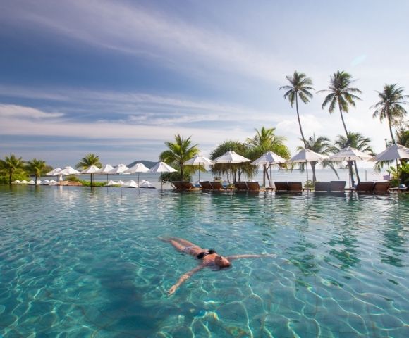 A person floats peacefully in a serene, turquoise pool surrounded by palm trees and white umbrellas under a clear sky, evoking a sense of relaxation.