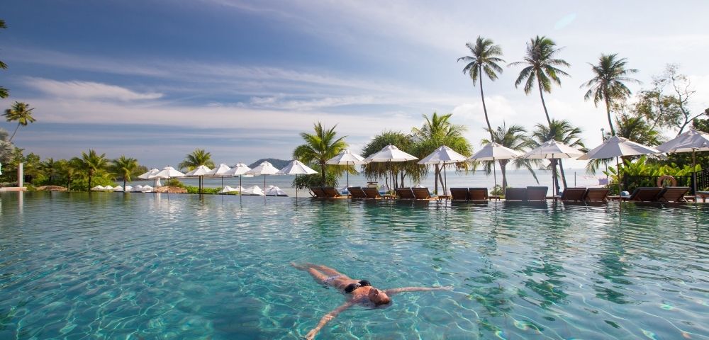A person floats peacefully in a serene, turquoise pool surrounded by palm trees and white umbrellas under a clear sky, evoking a sense of relaxation.
