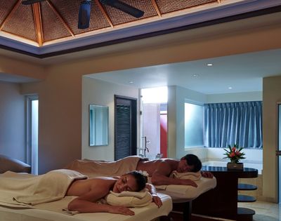Couple enjoys a relaxing massage on two spa tables in a serene room with soft lighting, a wooden ceiling, and a flower arrangemenCouple enjoys a relaxing massage on two spa tables in a serene room with soft lighting, a wooden ceiling, and a flower arrangement, conveying tranquility.t, conveying tranquility.
