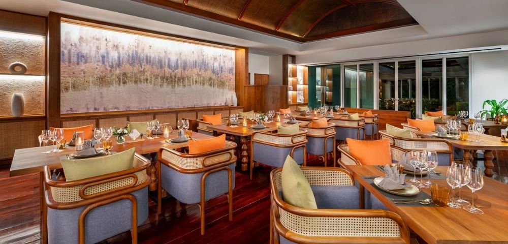 Elegant restaurant interior with wooden floors, beige and orange cushioned chairs, and tables set with wine glasses. Warm, inviting ambiance.