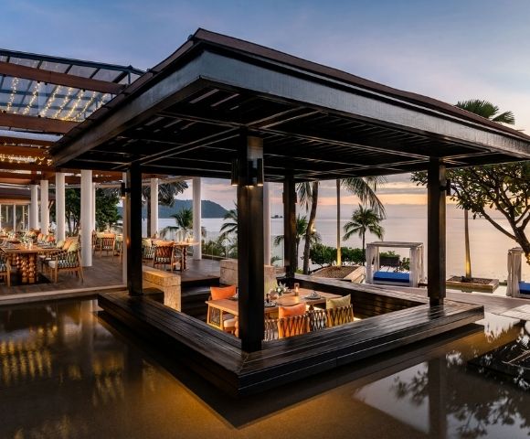 Outdoor restaurant with a modern wooden pavilion, overlooking the ocean at sunset. Warm lighting and palm trees create a serene and inviting atmosphere.