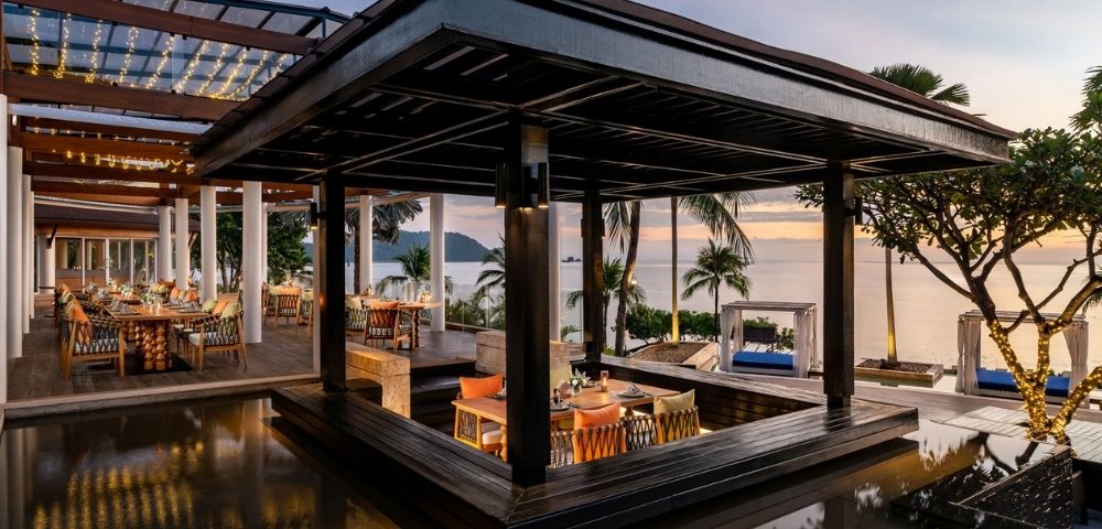 Outdoor restaurant with a modern wooden pavilion, overlooking the ocean at sunset. Warm lighting and palm trees create a serene and inviting atmosphere.