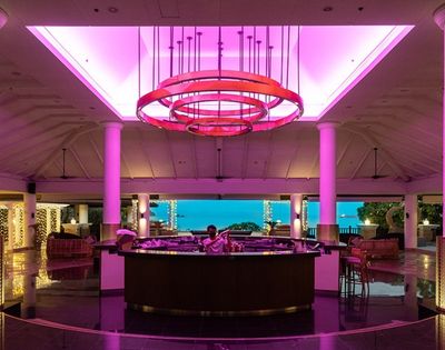 A modern lobby bar with vibrant pink ceiling lights, white pillars, and a view of the ocean. The atmosphere is elegant and inviting.