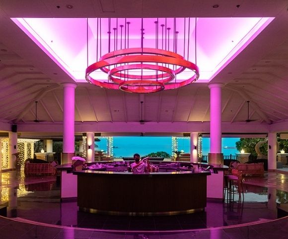 A modern lobby bar with vibrant pink ceiling lights, white pillars, and a view of the ocean. The atmosphere is elegant and inviting.