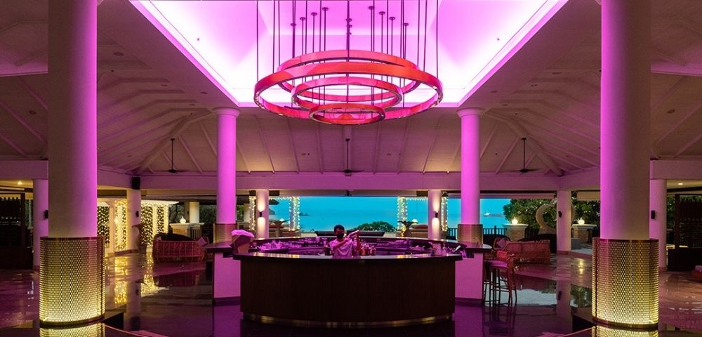 A modern lobby bar with vibrant pink ceiling lights, white pillars, and a view of the ocean. The atmosphere is elegant and inviting.