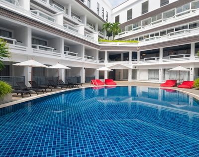 Outdoor hotel pool surrounded by white balconies, blue tiled water, black lounge chairs, red cushions, and white umbrellas under a clear sky. Relaxing atmosphere.
