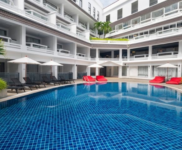 Outdoor hotel pool surrounded by white balconies, blue tiled water, black lounge chairs, red cushions, and white umbrellas under a clear sky. Relaxing atmosphere.
