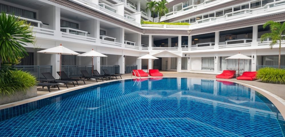 Outdoor hotel pool surrounded by white balconies, blue tiled water, black lounge chairs, red cushions, and white umbrellas under a clear sky. Relaxing atmosphere.