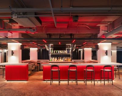 A modern bar with a vibrant red counter, black high stools, and bottles displayed against a mirrored backdrop. Neon lighting creates a trendy ambiance.
