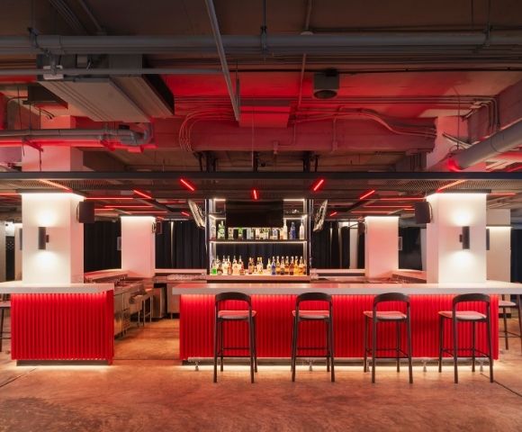 A modern bar with a vibrant red counter, black high stools, and bottles displayed against a mirrored backdrop. Neon lighting creates a trendy ambiance.