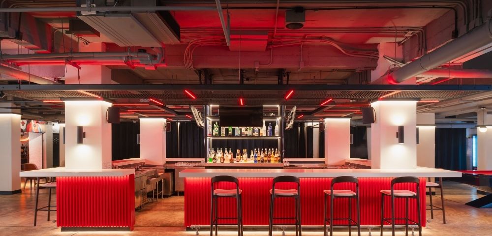 A modern bar with a vibrant red counter, black high stools, and bottles displayed against a mirrored backdrop. Neon lighting creates a trendy ambiance.