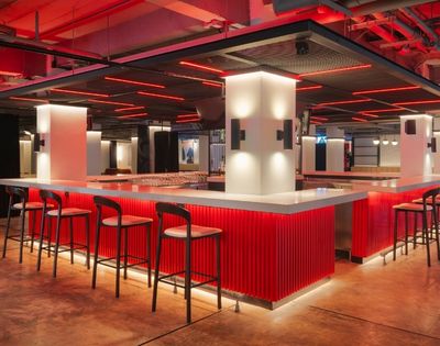 Modern bar interior with red lighting and sleek design. Features a white countertop, black chairs, and glowing accents, creating a stylish atmosphere.
