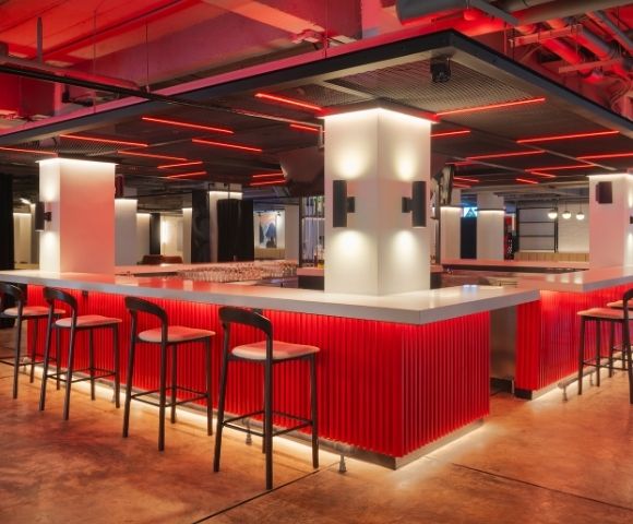 Modern bar interior with red lighting and sleek design. Features a white countertop, black chairs, and glowing accents, creating a stylish atmosphere.