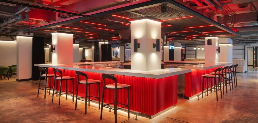 Modern bar interior with red lighting and sleek design. Features a white countertop, black chairs, and glowing accents, creating a stylish atmosphere.
