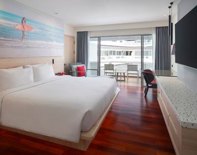 Modern hotel room with a large white bed, wooden flooring, wall art of a surfer, large TV, and a balcony with chairs. Bright and inviting atmosphere.