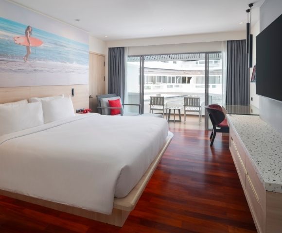 Modern hotel room with a large white bed, wooden flooring, wall art of a surfer, large TV, and a balcony with chairs. Bright and inviting atmosphere.