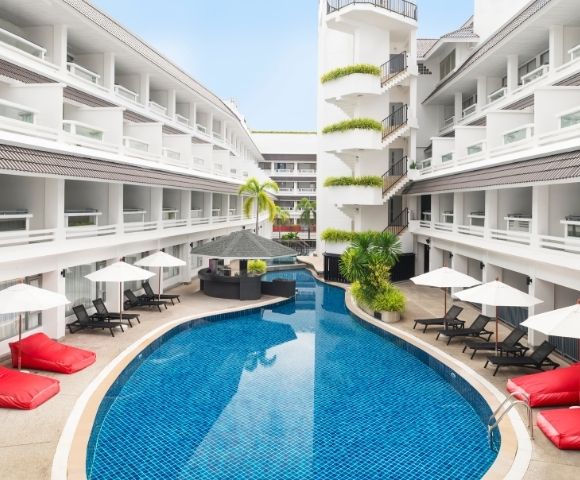 A serene hotel courtyard with a curved blue swimming pool, surrounded by white balconies. Red lounge chairA serene hotel courtyard with a curved blue swimming pool, surrounded by white balconies. Red lounge chairs and white umbrellas line the poolside.s and white umbrellas line the poolside.
