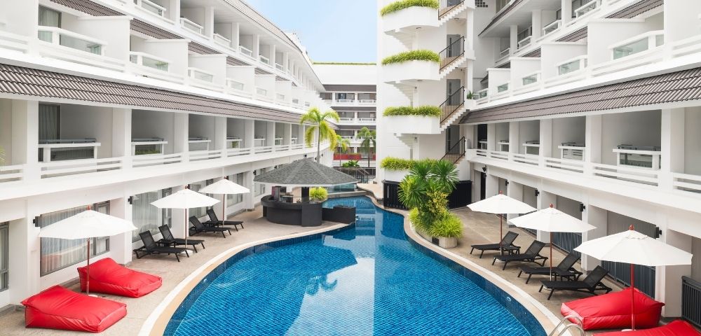 A serene hotel courtyard with a curved blue swimming pool, surrounded by white balconies. Red lounge chairs and white umbrellas line the poolside.