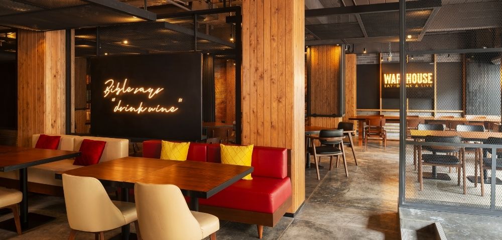 Modern cafe interior with warm lighting, wooden accents, and red cushioned seating. Neon signs read 