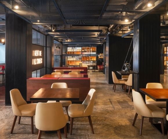 Modern restaurant interior with warm lighting, wooden tables, beige cushioned chairs, and decorative shelves. Cozy and inviting atmosphere.