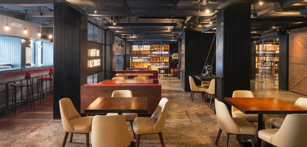 Modern restaurant interior with warm lighting, wooden tables, beige cushioned chairs, and decorative shelves. Cozy and inviting atmosphere.