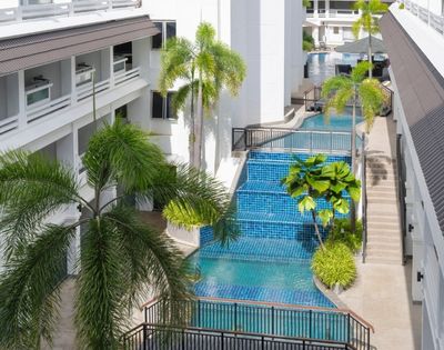 A modern hotel courtyard features a serene blue-tiled pool flanked by lush palm trees. The atmosphere is tranquil and inviting, with bright, natural light.