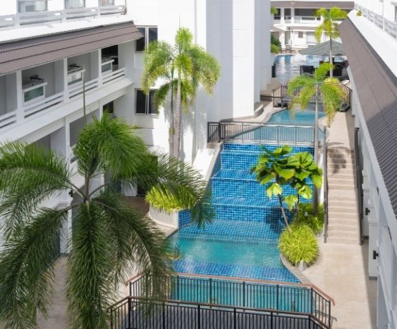 A modern hotel courtyard features a serene blue-tiled pool flanked by lush palm trees. The atmosphere is tranquil and inviting, with bright, natural light.