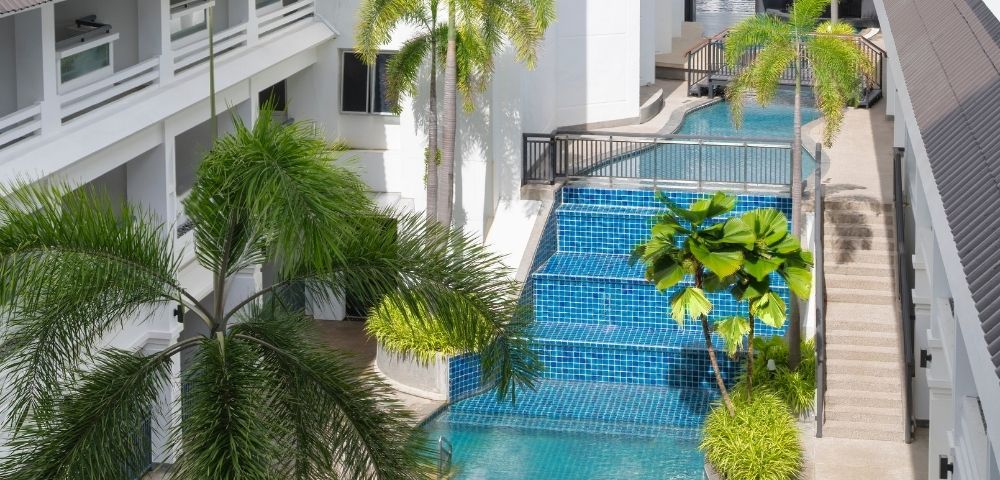 A modern hotel courtyard features a serene blue-tiled pool flanked by lush palm trees. The atmosphere is tranquil and inviting, with bright, natural light.