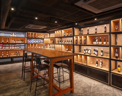Cozy wine cellar with dim lighting, wooden shelves lined with wine bottles, and a tall wooden table with bar stools, creating a warm, inviting atmosphere.