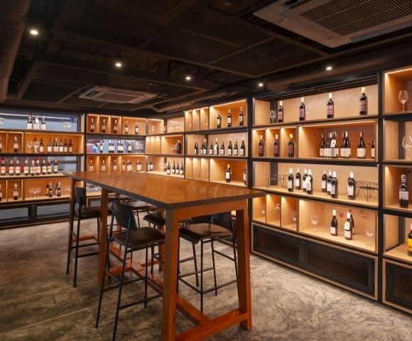 Cozy wine cellar with dim lighting, wooden shelves lined with wine bottles, and a tall wooden table with bar stools, creating a warm, inviting atmosphere.