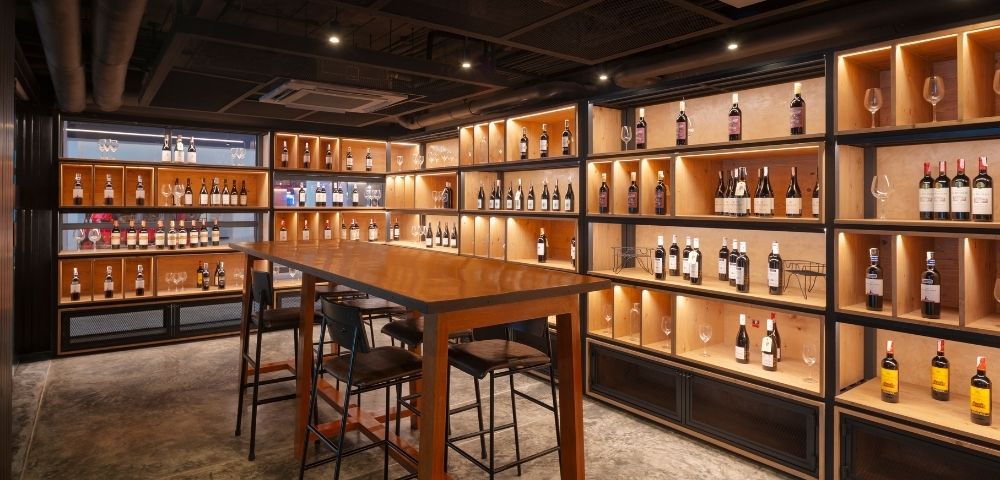 Cozy wine cellar with dim lighting, wooden shelves lined with wine bottles, and a tall wooden table with bar stools, creating a warm, inviting atmosphere.
