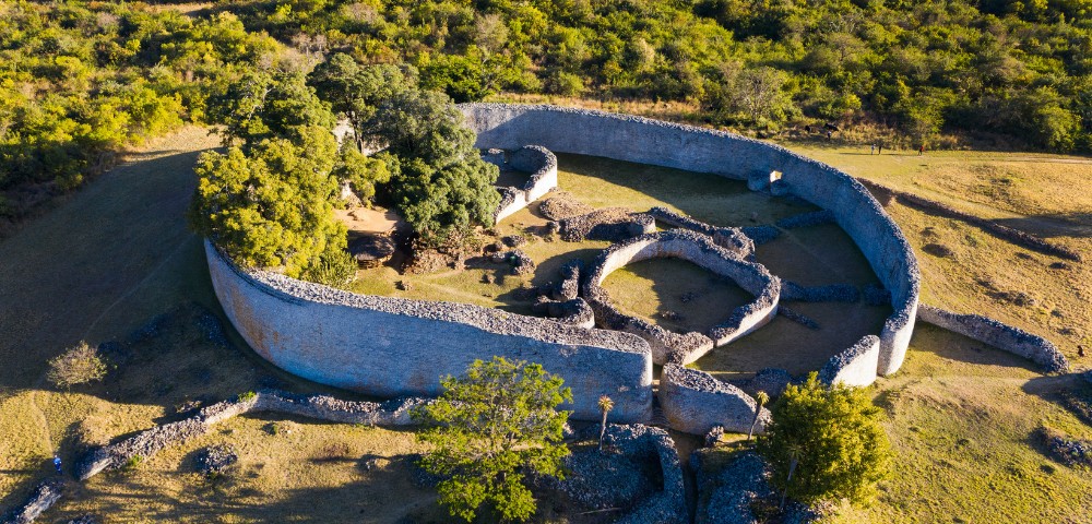 Aerial view of ancient circular stone ruins surrounded by green trees and hillside vegetation under sunlight.